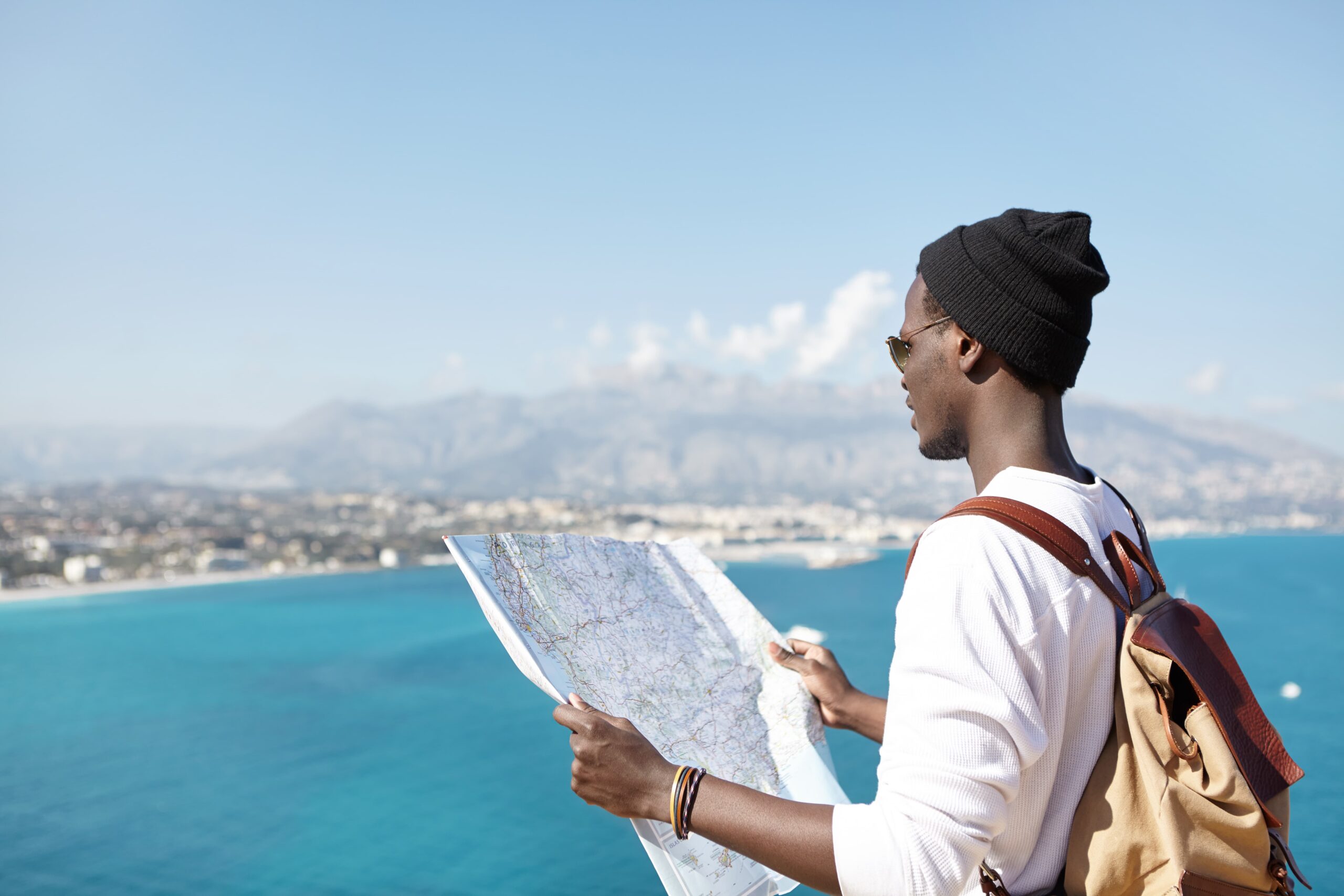 fashionable-young-male-backpacker-holding-paper-map-standing-top-mountain-ocean