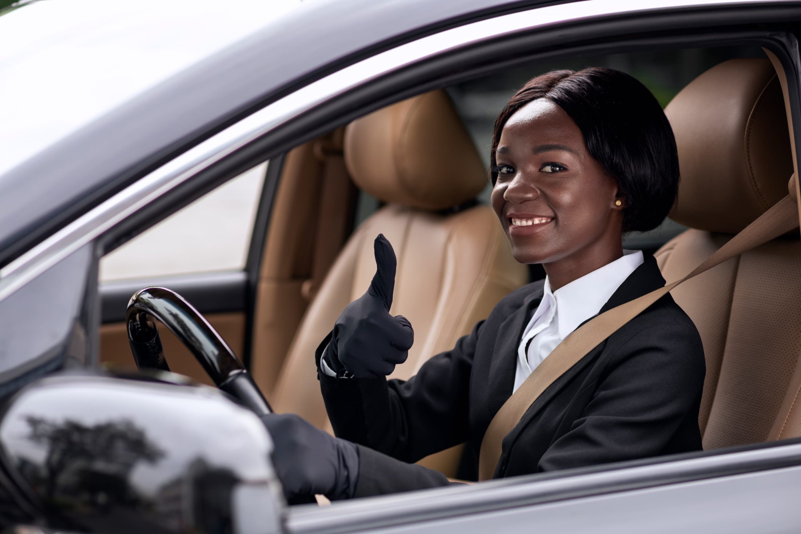 Person in formal attire with black suit and white shirt sitting in the driver’s seat of a car, wearing a seatbelt and gloves, smiling and giving a thumbs-up gesture, with beige leather seats and a black steering wheel visible.