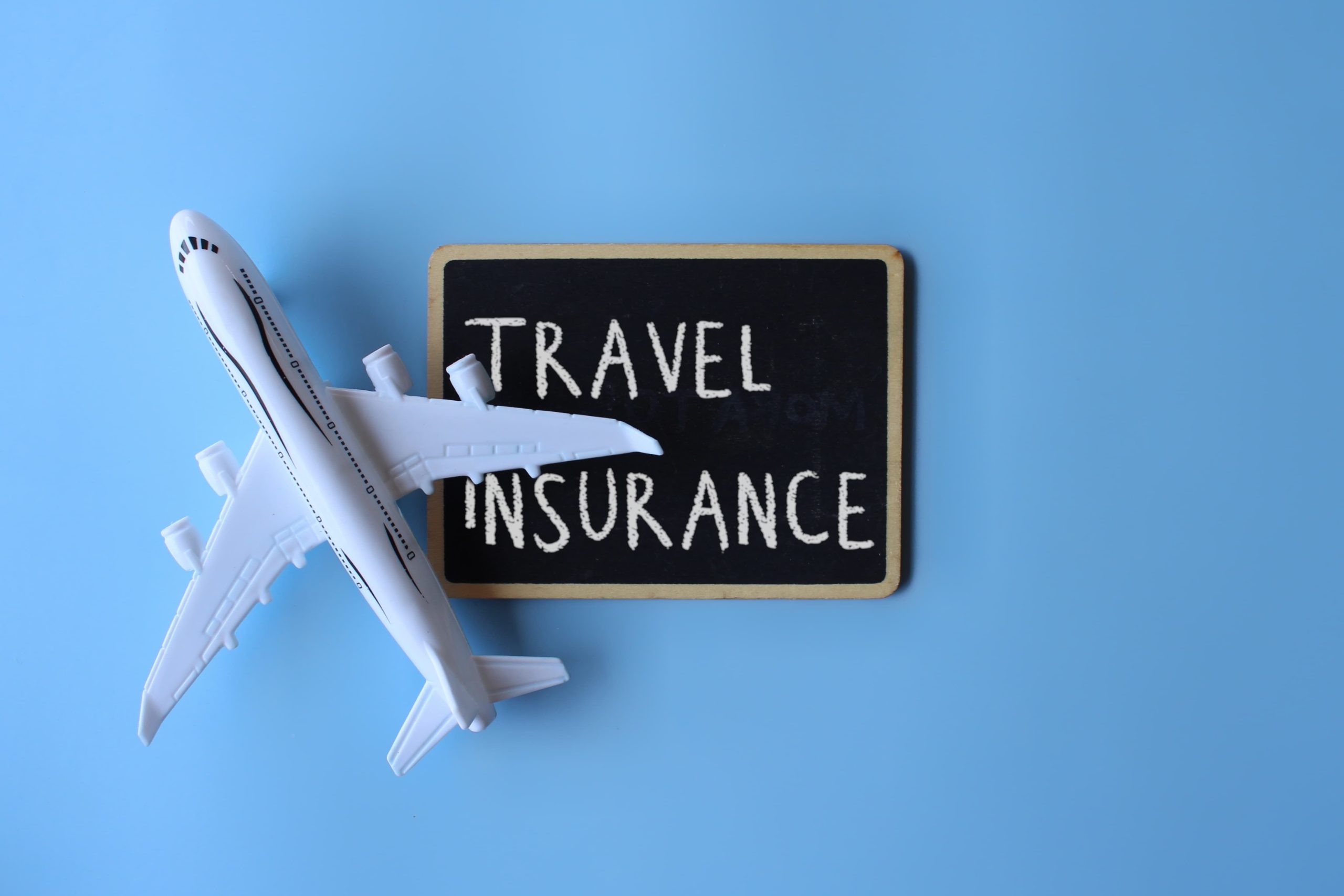 Model airplane placed beside a small blackboard with 'TRAVEL INSURANCE' written in white chalk on a light blue background, symbolizing travel protection.